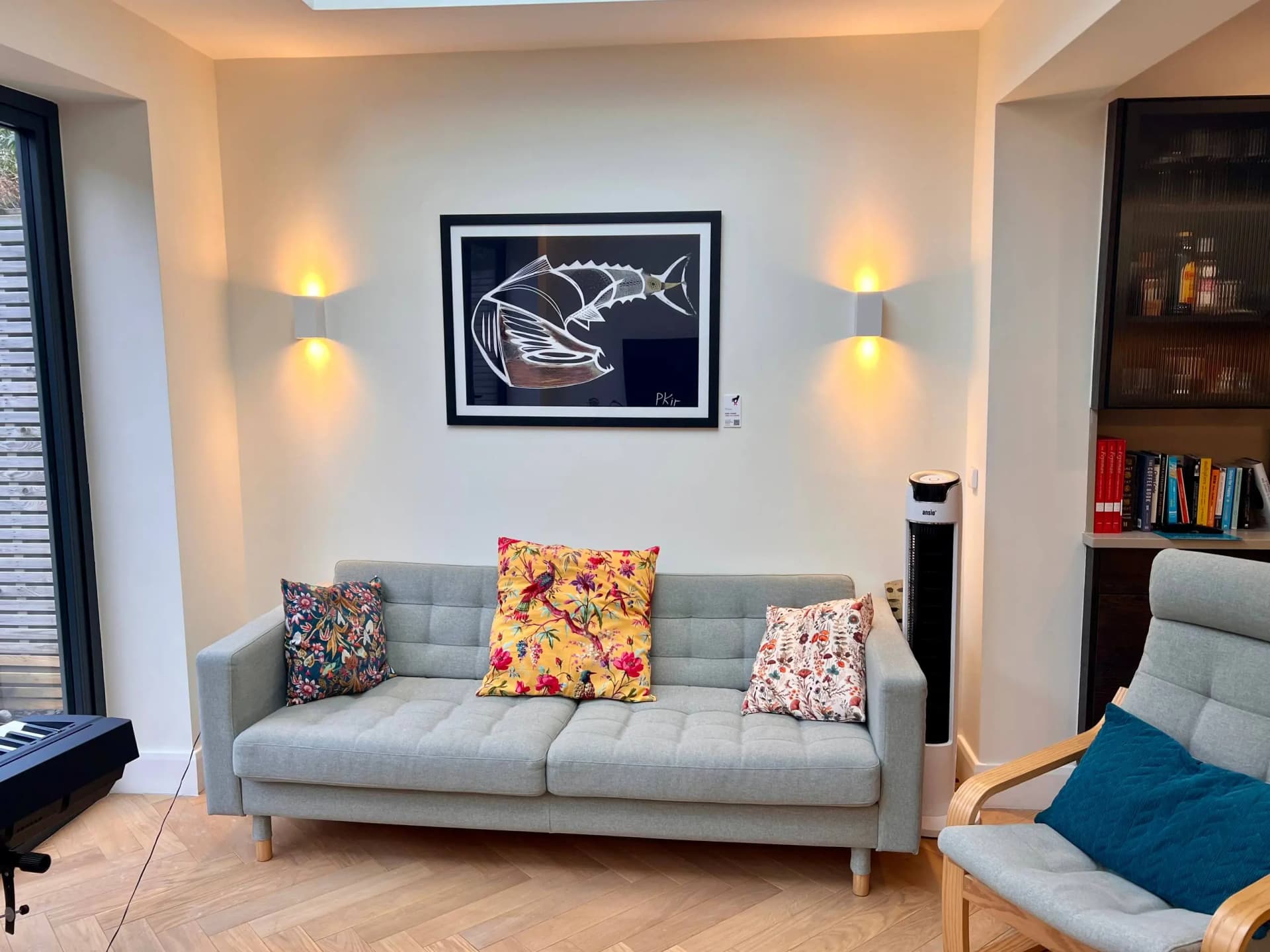 Grey sofa with patterned cushions beneath a framed black-and-white fish artwork between two warm wall lights