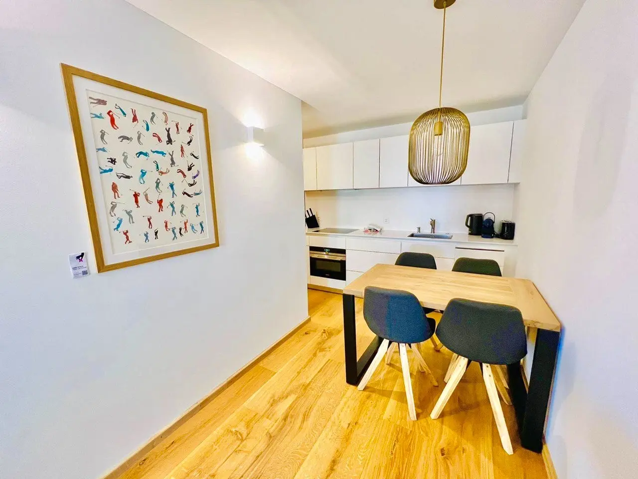 Small holiday let dining area with a wooden table and dark chairs beside a framed artwork of colourful golfers