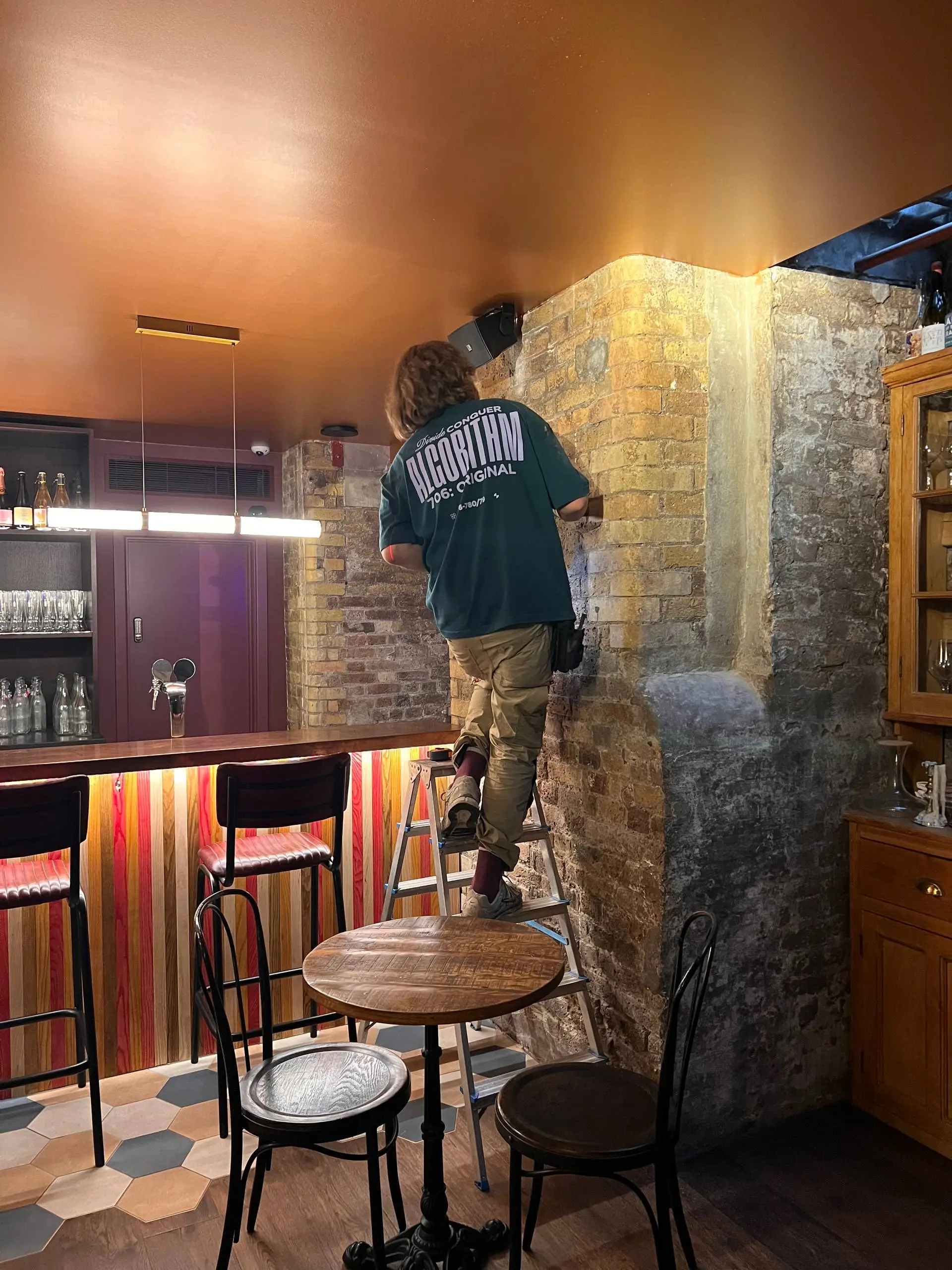 Installer on a stepladder fitting a hanging rail to an exposed brick wall beside the bar