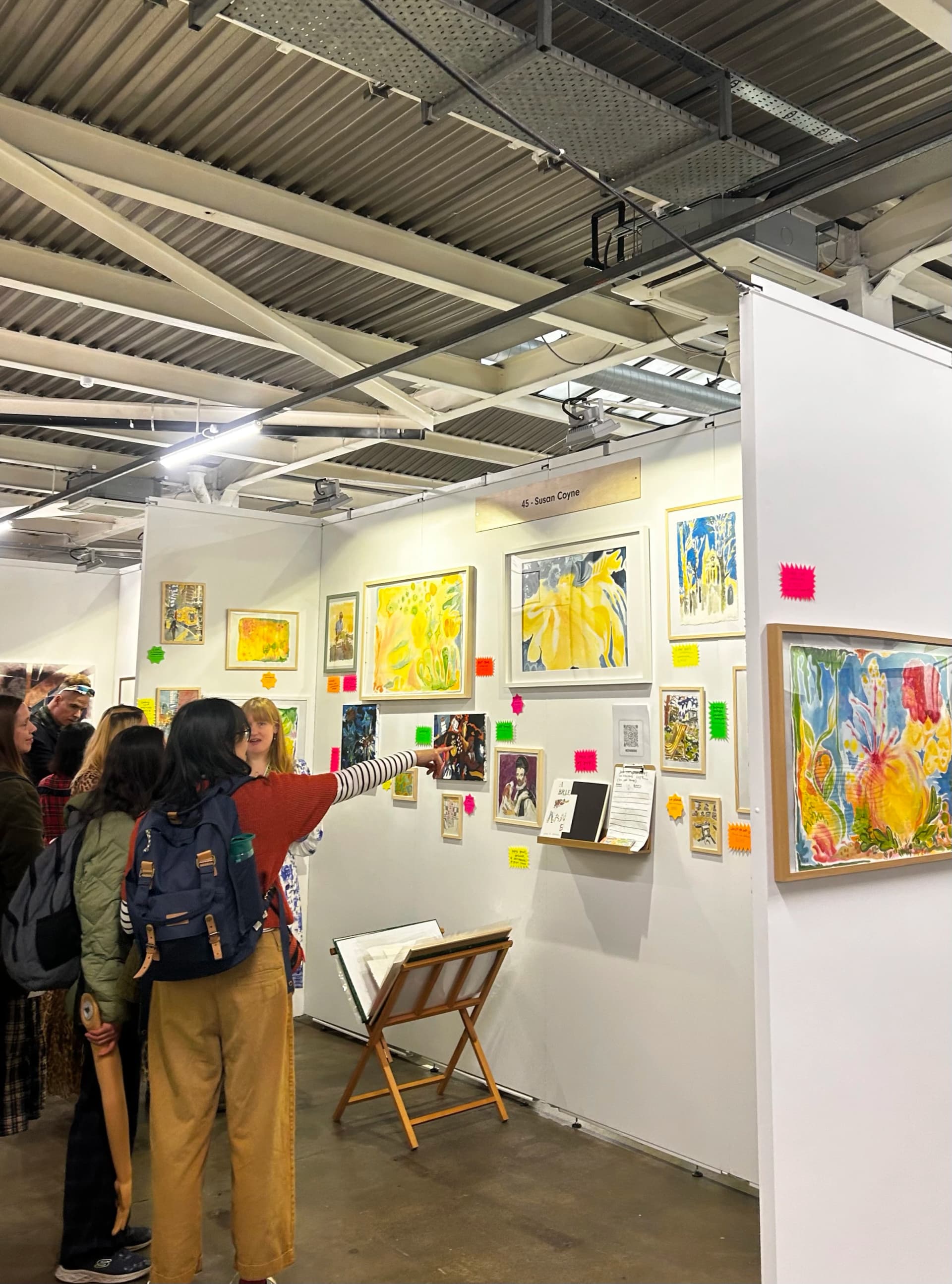 Independent artist exhibiting artwork at a London art fair, with visitors looking at framed works on a stand